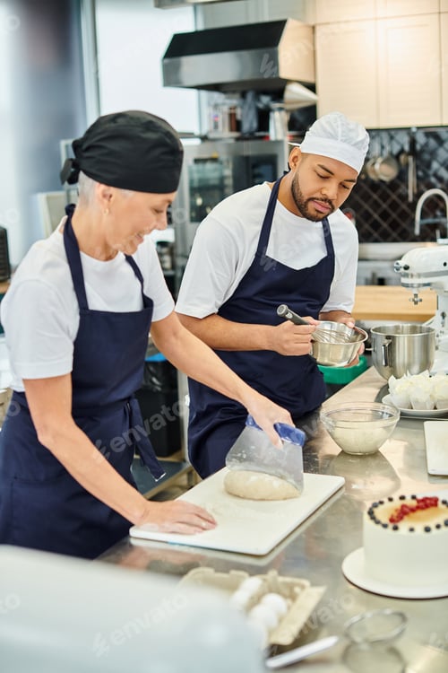 Preview: focus on young african american chef in toque whisking next to his blurred jolly mature colleague
