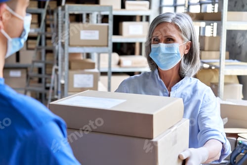 Preview: Female manager giving parcels boxes to courier wearing face masks in warehouse.