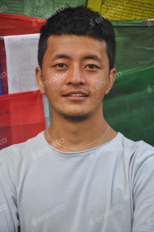 Preview: Close up of a Ladakhi young guy looking at camera, standing against Buddhist prayer flags