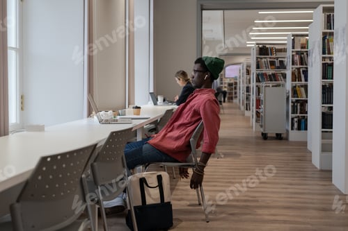Preview: Tired Black student man sitting at desk with laptop in university library, looking educational video