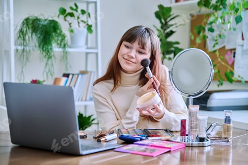 Preview: Young female showing cosmetics to laptop webcam, sitting at table at home