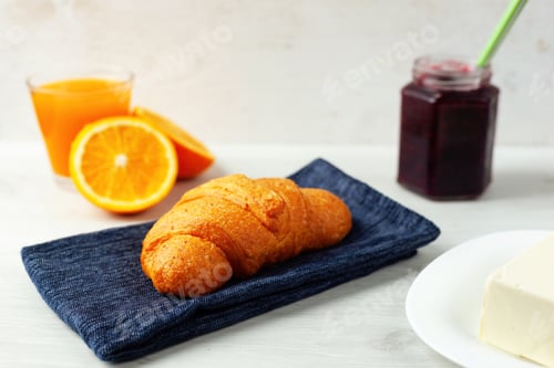 Preview: Delicious croissant resting on blue napkin with orange juice, jam and butter for breakfast