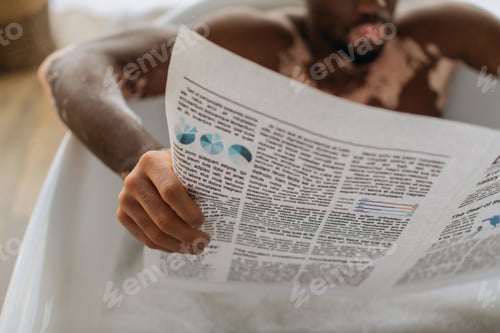 Preview: Cropped view of blurred african american man with vitiligo reading newspaper while taking bath in