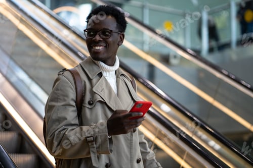 Preview: Smiling Black traveler man stands on escalator in airport terminal or railway station, using mobile