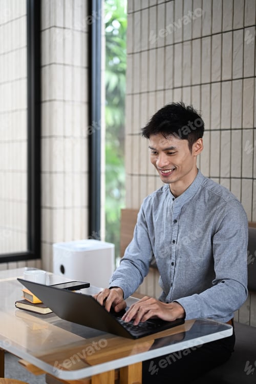 Preview: Smiling young man working on laptop at a modern cafe with books and coffee on the table