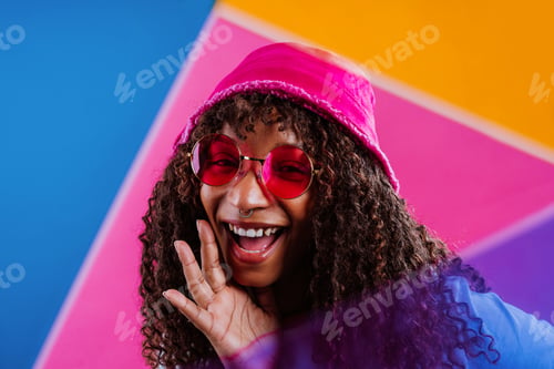 Preview: Young woman wearing pink sunglasses and beanie smiling on colorful background