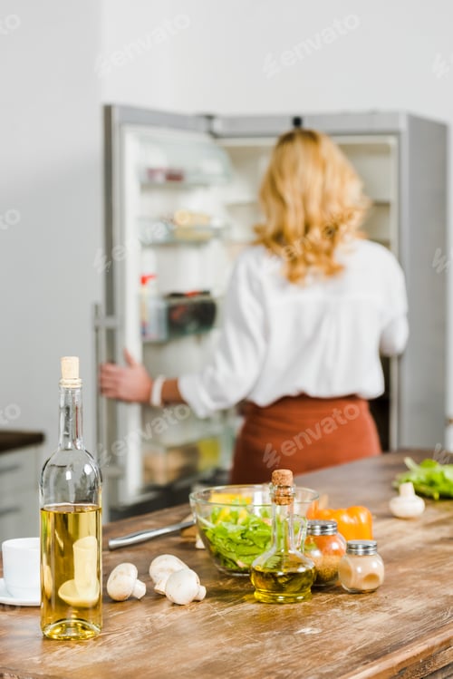 Preview: selective focus of mature woman taking something from fridge in kitchen, vegetables and wine on