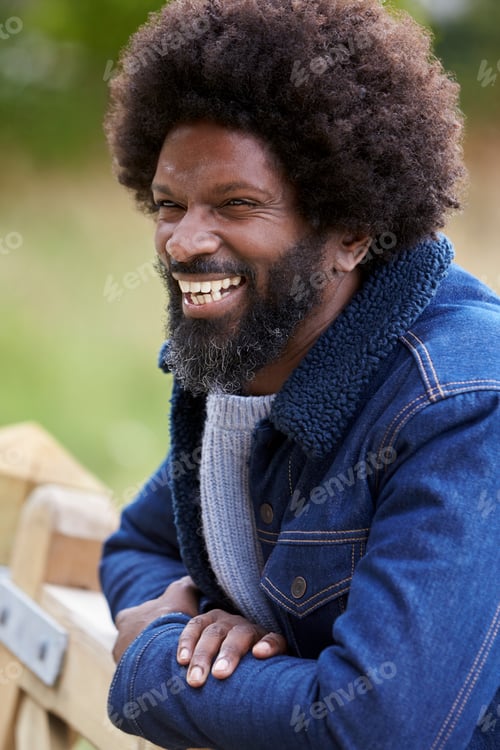 Preview: Black man leaning on a wooden fence in the countryside laughing, close up