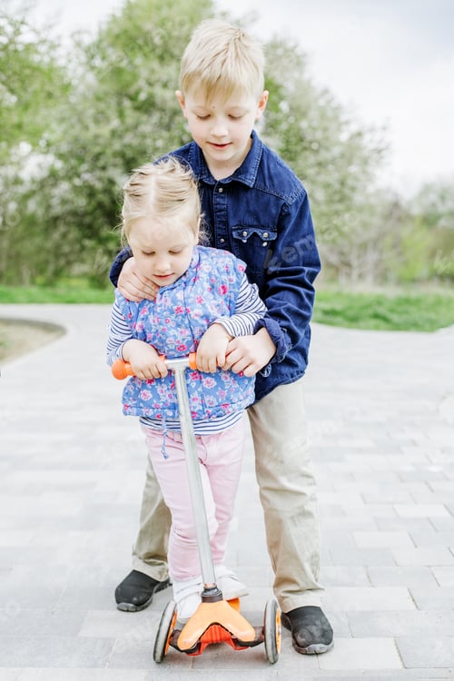 Preview: Child boy teaches little sister how to ride a children's scooter. Active rest on playground.