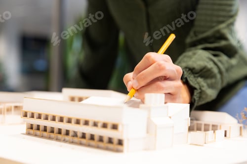 Preview: Midsection of caucasian female architect examining model with pencil at modern workplace