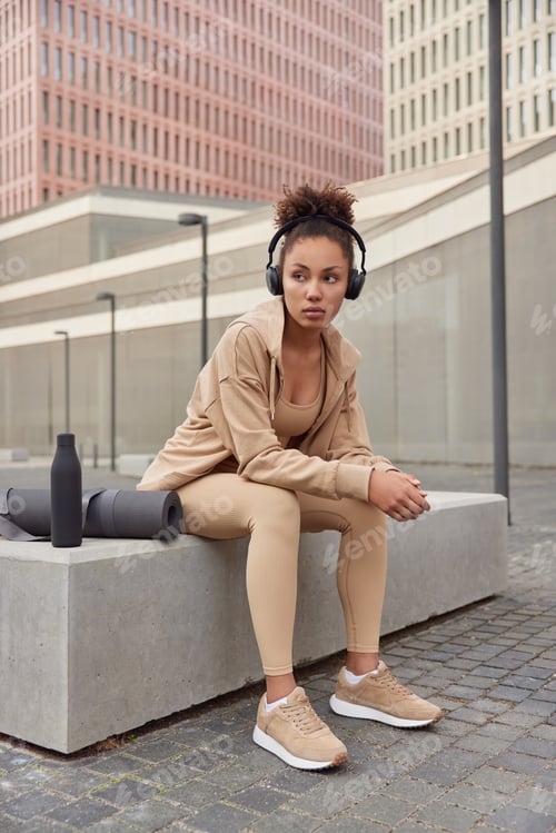 Preview: Sporty tired woman dressed in sportswear listens music from playlist via headphones poses outdoors n