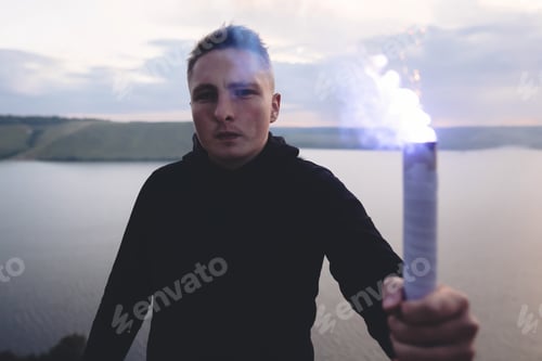 Preview: Ultras hooligan holding blue flare torch in hand, standing on top of rock mountain