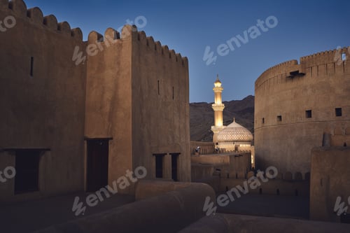 Preview: Evening view between watchtowers of historic fort to illuminated mosque. Nizwa, Sultanate of Oman.