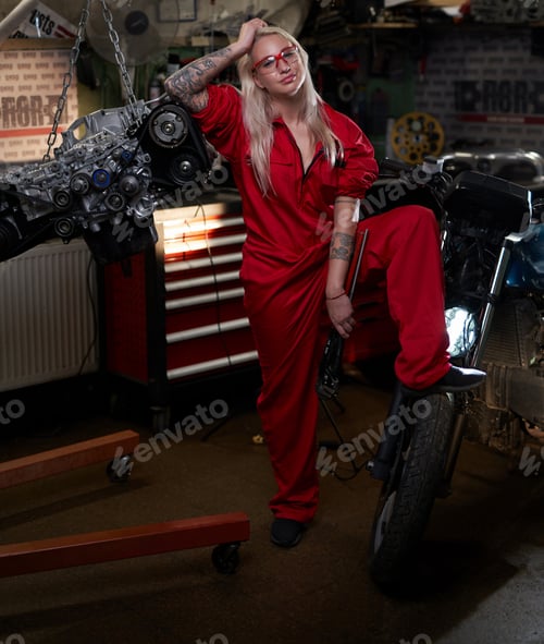 Preview: Blond female mechanic with tattooed hands in orange overalls stands in garage or workshop