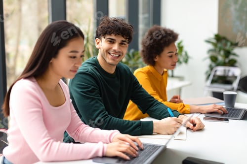 Preview: Group of diverse international students sitting at desk in coworking space, focus on smiling guy