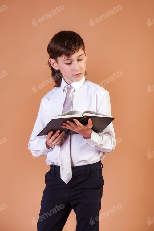Preview: Portrait of pensive 8 year old schoolboy reading book at beige backdrop, intelligent smart look