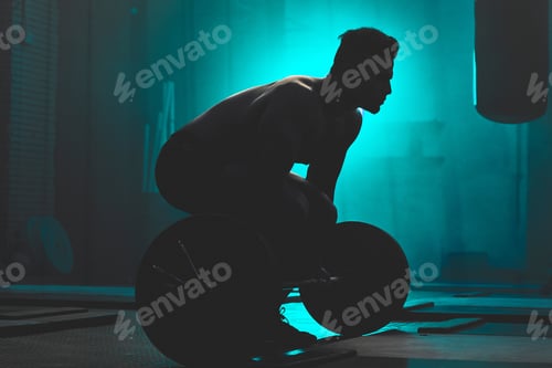 Preview: Silhouette of an unrecognizable boxer man lifting weights in a dark gym. Male athlete working out i