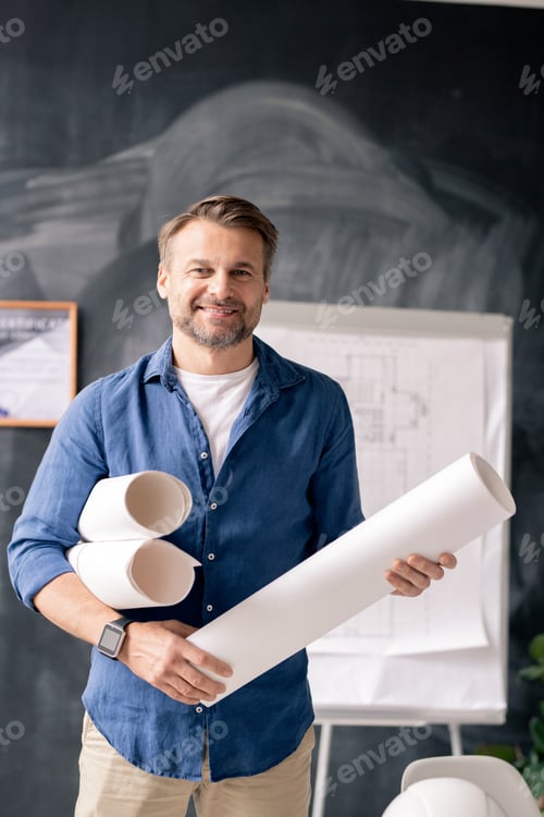Preview: Successful mature engineer with rolled blueprints standing in front of camera