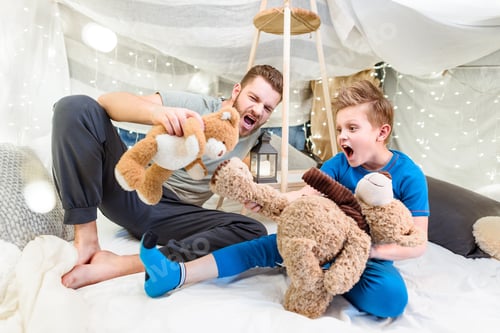 Preview: Excited father and son sitting in blanket fort and playing with teddy bears