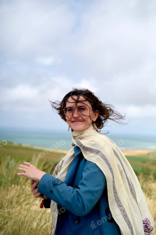 Preview: Woman with glasses smiling in a windy field with the sea in the background.