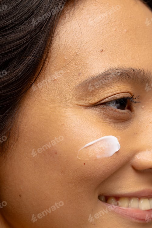 Preview: Close up of smiling asian woman with dark hair with skin cream on her cheek