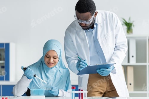 Preview: African american scientist in safety goggles writing on clipboard near smiling arabian colleague