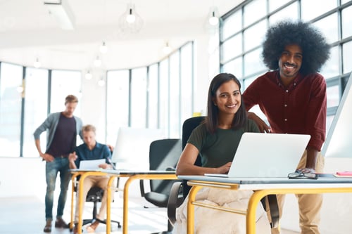 Preview: Portrait of two happy designers posing at a desk while their colleagues work in the background