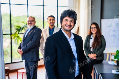 Preview: Confident Indian business leader standing with folded hands, colleagues out of focus in the office