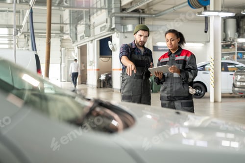 Preview: Young repairman in uniform pointing at front part of car while describing it