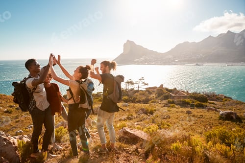 Preview: Young adult friends on a hike celebrate reaching a summit near the coast, full length, side view