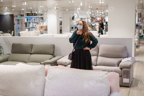 Preview: Young pensive woman in face mask standing in front of couch in furniture store department