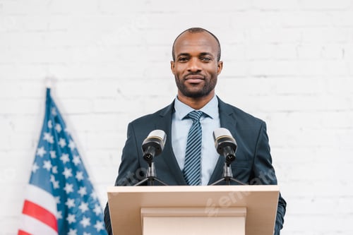 Preview: happy african american speaker standing near microphones and american flag