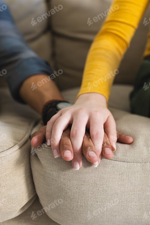 Preview: Diverse couple sitting on couch and holding hands in living room at home, copy space