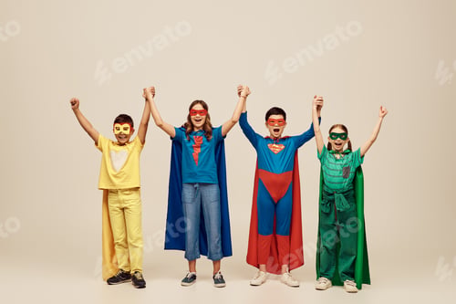 Preview: happy interracial kids in colorful superhero costumes with cloaks and masks raising and holding