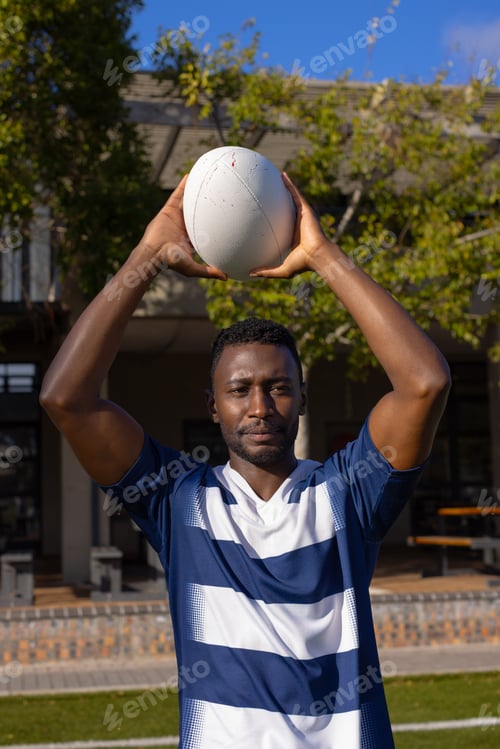 Preview: African American young male athlete holding rugby ball above head on field outdoors