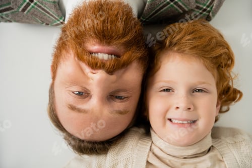 Preview: top view of redhead boy with bearded dad smiling at camera while lying on grey background