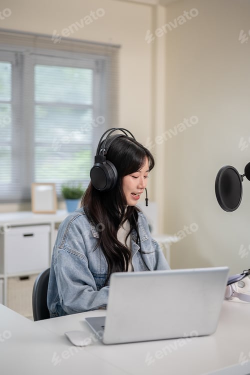 Preview: Asian woman looking at clipboard while hosting radio or podcast through laptop with headset and mic.