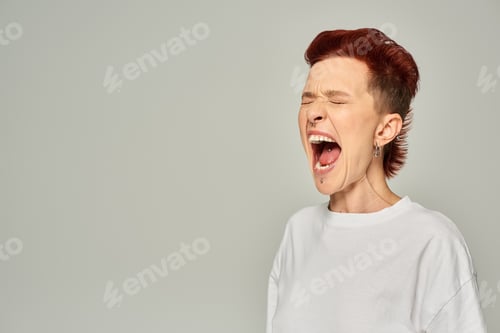 Preview: emotional queer person in white t-shirt standing and screaming with closed eyes on grey backdrop