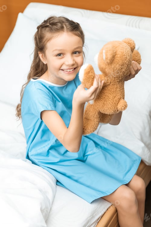 Preview: Little patient with teddy bear sitting on bed in hospital