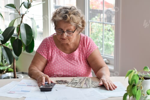 Preview: Senior woman sitting at desk using calculator to count money to pay bills