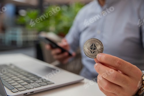 Preview: Ethereum coin held in the hand of a trader monitoring the market with his laptop and smartphone.