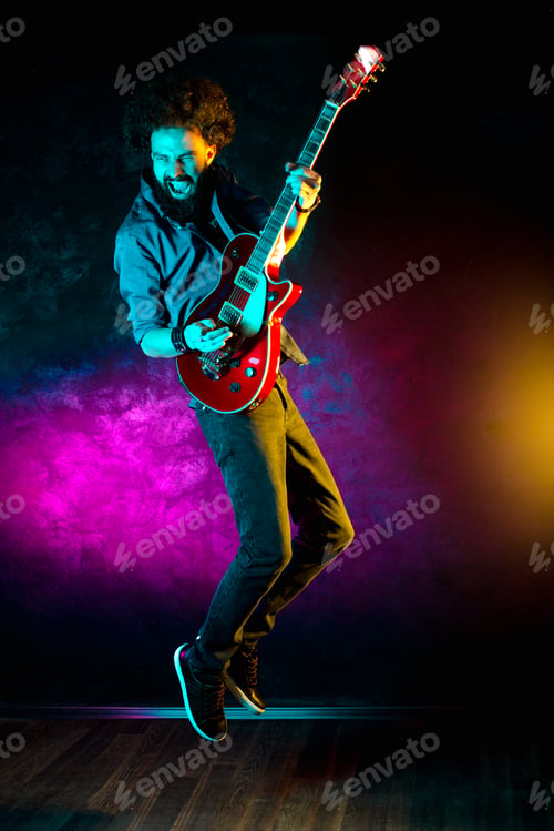 Preview: Young hipster man with curly hair jump with red guitar in neon lights.