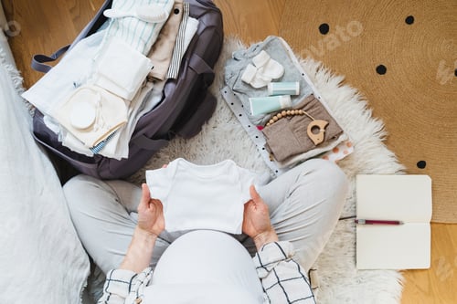 Preview: Pregnant woman holding baby bodysuit and packing maternity hospital bag.