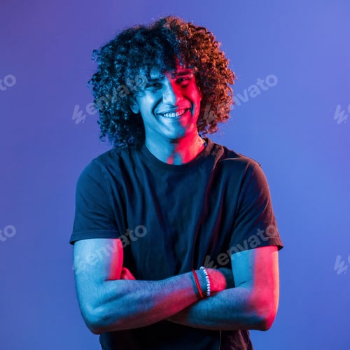 Preview: Posing for a camera. Young beautiful man with curly hair is indoors in the studio with neon lighting