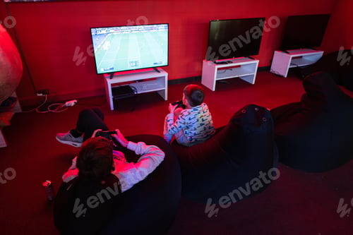 Preview: Two boys gamers play football gamepad video game console in red gaming room.