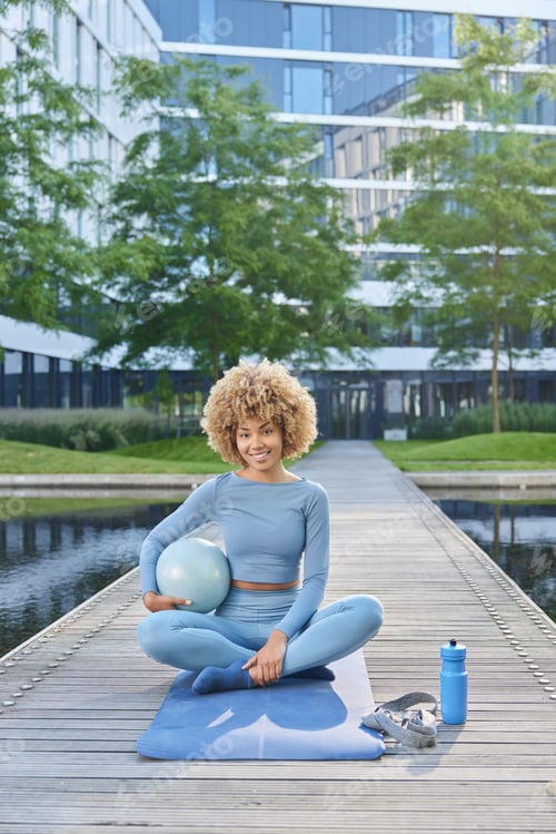 Preview: Vertical shot of active slim woman in blue sportswear sits crossed legs with ball practices yoga or