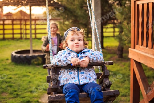 Preview: Cute boy in hat and suit riding swing on backyard and laughing. Sister playing with little brother
