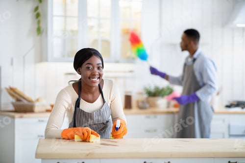 Preview: Happy African American woman wiping kitchen table while her husband dusting on background, copy