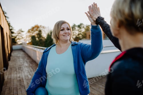 Preview: Happy overweight woman high fiving with personal trainer outdoors on gym terrace