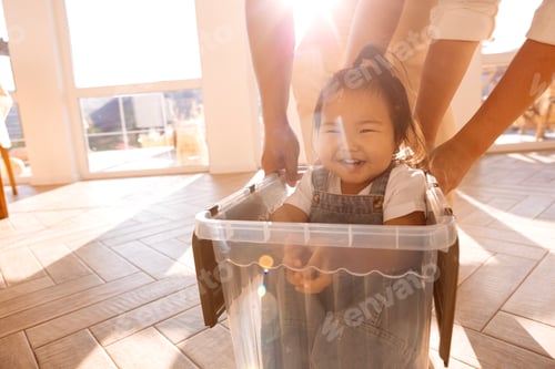 Preview: Young laughing Asian parents roll their daughter in a plastic box around the house